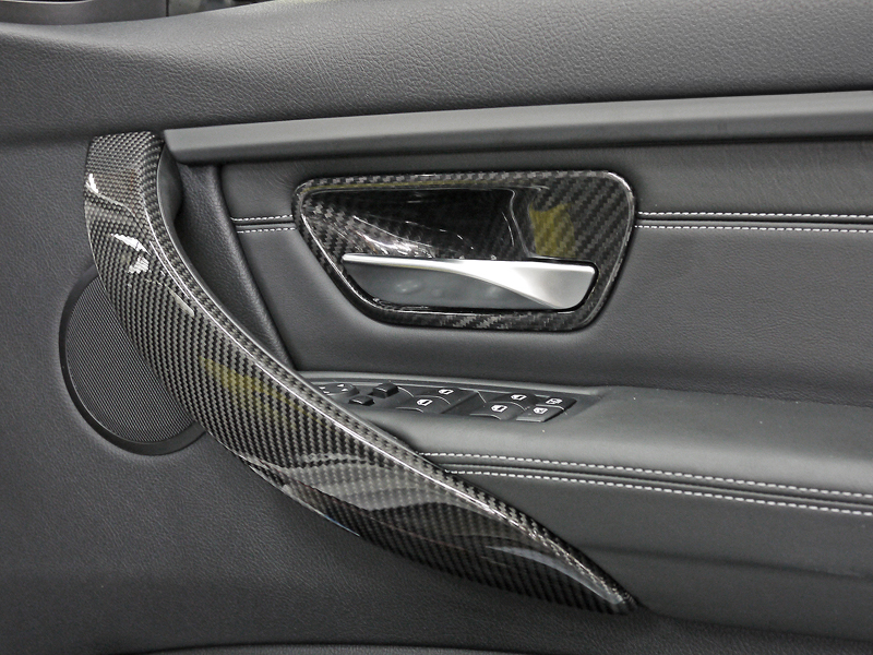 Close-up image of a car's interior door handle trim made of dry carbon fiber, installed on a vehicle's door panel.