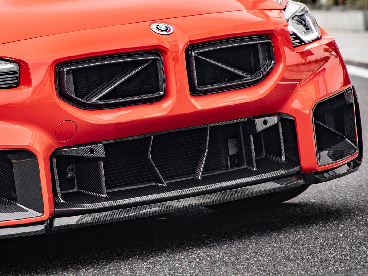 Close-up of a red BMW car's front bumper featuring the AutoTecknic dry carbon fiber OEM-Spec center bumper trim.