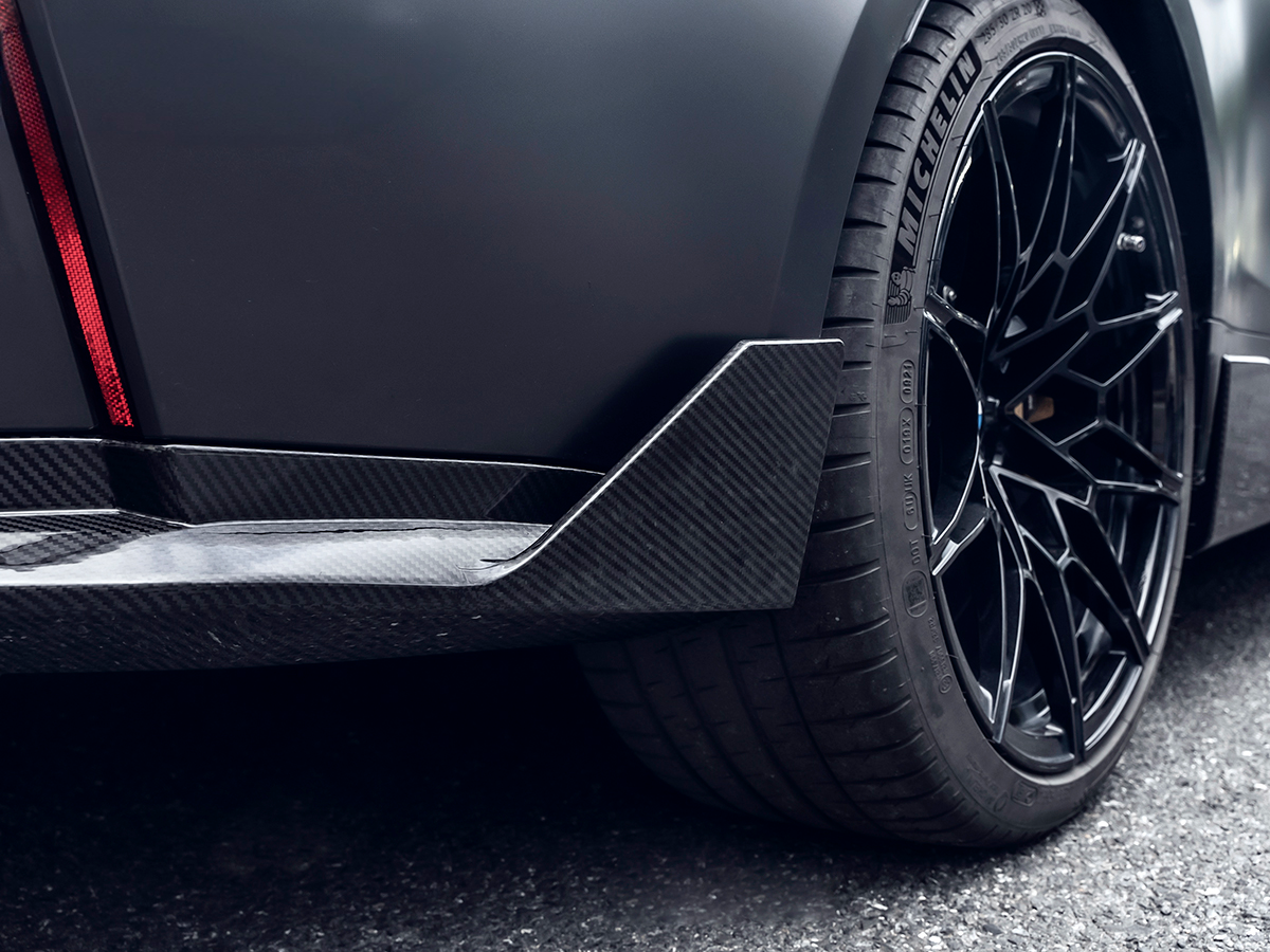 Close-up of a black car's rear side splitter and wheel, showcasing the AutoTecknic dry carbon fiber product.
