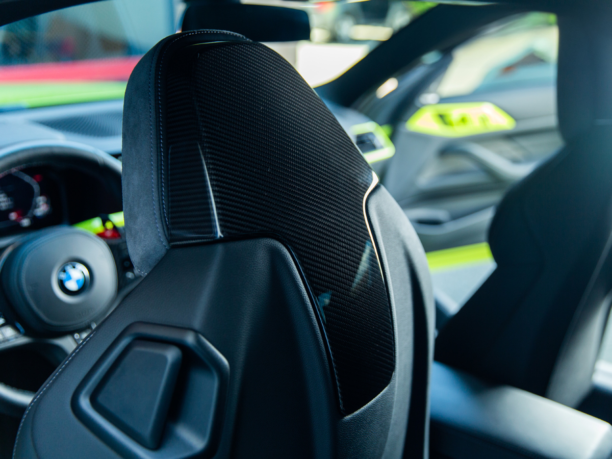 Close-up image of a black dry carbon seat back cover installed inside a vehicle's interior, focusing on texture and gloss.