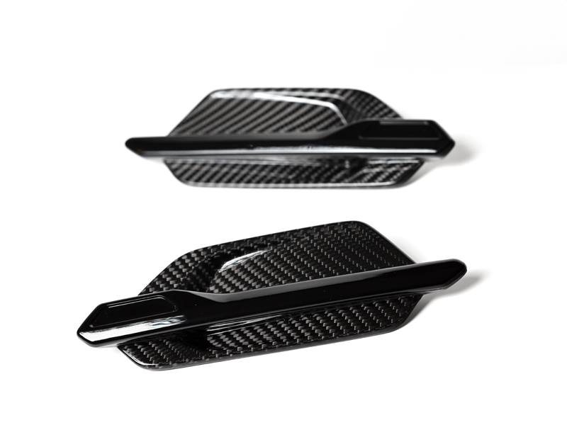 A pair of AutoTecknic dry carbon fiber fender trims, designed for a BMW M2, displayed against a white background.