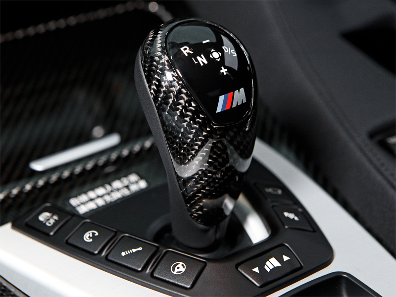 Close-up image of a carbon fiber gear selector cover with a BMW logo, situated inside a vehicle's interior.