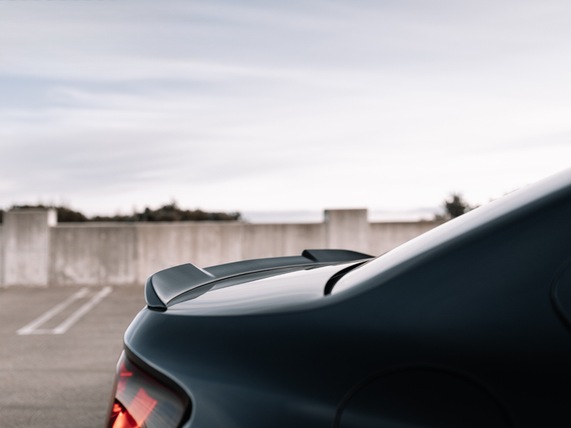 A close-up image of a black AutoTecknic dry carbon fiber competition trunk spoiler installed on the rear of a car.