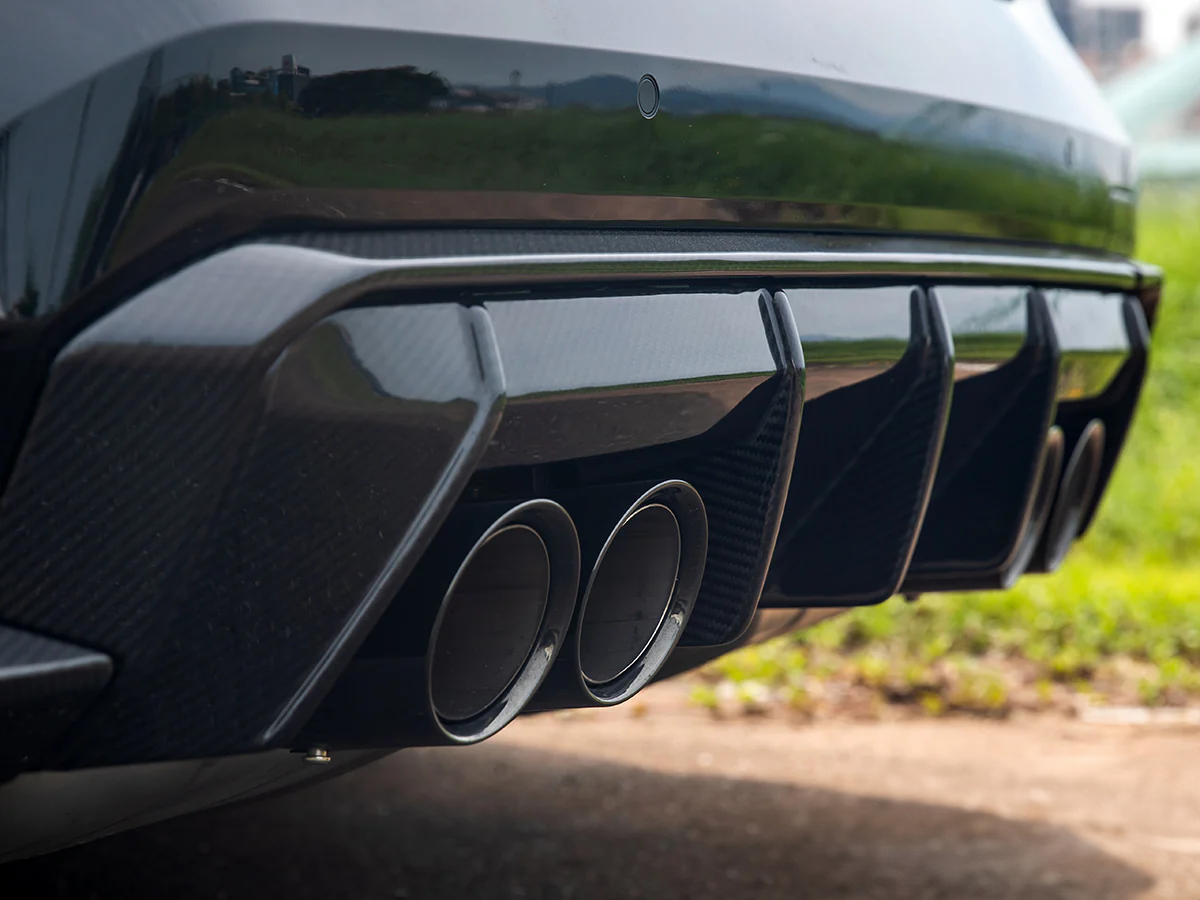 Close-up image of a black carbon fiber rear diffuser trim set installed on the rear bumper of a vehicle.