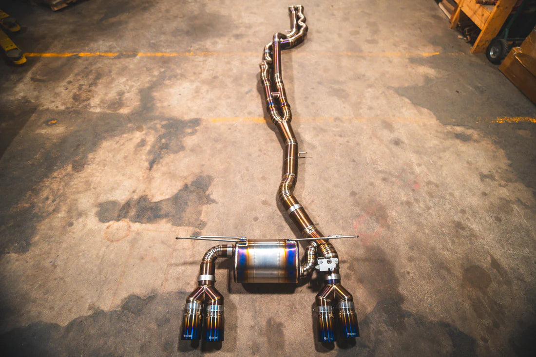 A performance exhaust system for BMW M3 and M4, featuring dual mufflers and electronic exhaust valves, laid on a concrete floor.