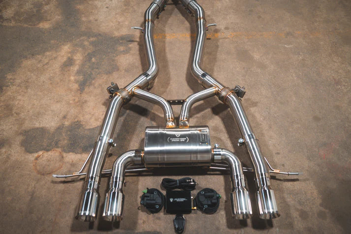 A performance exhaust system for a BMW M2, featuring stainless steel pipes andValved components, laid out on a concrete floor.