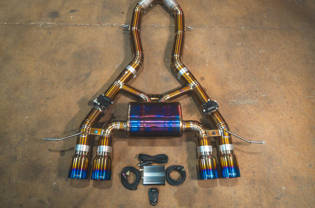 A performance exhaust system for BMW M3 and M4, featuring a stainless steel or titanium construction with blue and silver colors, including multiple exhaust pipes and valves.