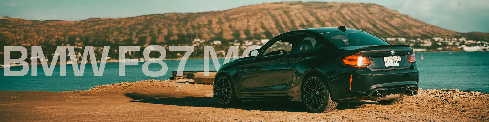Green BMW F87 M2 car parked by a body of water with mountains in the background